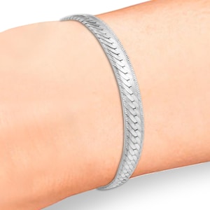 Puede incluir: Un brazalete plateado con un diseño de espiga. El brazalete se lleva en la muñeca y tiene un acabado liso y pulido. El diseño presenta una serie de eslabones entrelazados y angulados. El ancho es de aproximadamente 0,5 cm.