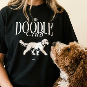 Puede incluir: Una camiseta negra de manga corta con el texto blanco THE DOODLE CLUB impreso en una fuente serif sobre una ilustración gráfica blanca de un perro caminando. El diseño está centrado en el pecho de la camiseta hecha de tela de algodón.
