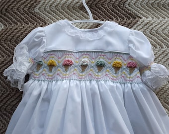 May include: White smocked baby dress with a lace collar and sleeves. The dress features a design of ice cream cones with colorful scoops.