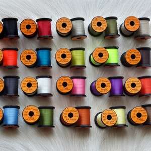 May include: A collection of 20 spools of Uni-Thread fly tying thread in various colors, including red, blue, green, black, white, and brown. Each spool is labeled with the thread color and yardage.