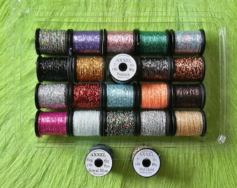 Könnte beinhalten: Eine transparente Plastikbox gefüllt mit 20 Spulen bunten, glitzernden Fadens. Jede Spule ist mit "AXXEL" beschriftet und hat eine andere Farbe, darunter Silber, Gold, Rot, Blau, Grün und Lila.