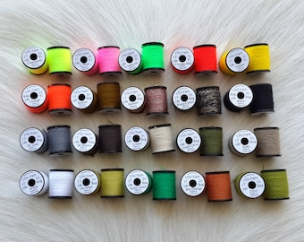 Könnte beinhalten: Eine Sammlung von 24 Garnrollen in verschiedenen Farben, darunter Schwarz, Weiß, Grün, Orange, Rot, Braun und Grau. Jede Rolle ist mit "UNI-Yarn" und der Länge des Garns in Yards gekennzeichnet.