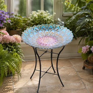 May include: A decorative glass bird bath with a peacock feather design in shades of blue, gold, and pink. The bird bath sits on a black metal stand. The bird bath is approximately 20 inches in diameter.