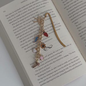May include: A gold-colored bookmark with a chain of charms, including a strawberry, a lightning bolt, a star, and a heart, rests on the open pages of a book. The book's pages are white with black text.
