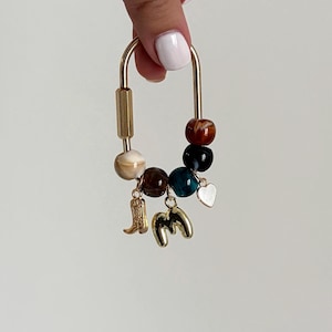 May include: A gold-toned metal carabiner keychain featuring a collection of round, multi-colored marbled beads in shades of brown, black, and teal. Three gold-toned charms hang from the beads, including a cowboy boot, the letter M, and a heart.
