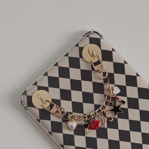 May include: A gold-toned phone charm with various charms, including a heart, key, lips, star, and letters, attached to a phone case with a black and white checkered pattern.