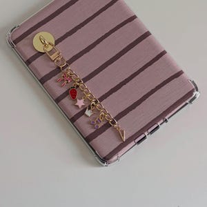 May include: A gold-colored phone charm with a chain and various charms, including a strawberry, star, and musical notes. The charm is attached to a phone case with a striped pattern in shades of mauve and brown.