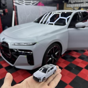 Op de afbeelding: Een miniatuur witte sedan-automodel wordt in een hand gehouden, met een grotere, full-size witte auto op de achtergrond. Het modelauto heeft een zwart dak en gedetailleerde kenmerken. De achtergrond omvat een rood-zwarte geblokte vloer en een winkelomgeving.