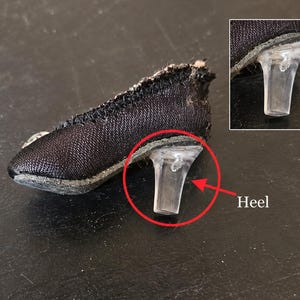 以下が含まれることがあります： 透明なプラスチック製のヒールが付いた黒い布製のミニチュアシューズ。靴は先が尖ったデザインで、縁に沿って目に見えるステッチが施され、アッパーは質感のある布地で作られています。赤い円がヒールを囲み、Heelという文字が添えられています。