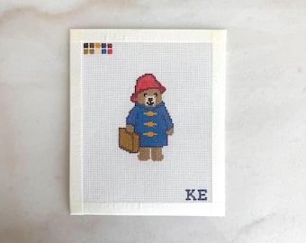 May include: A needlepoint canvas featuring a cross-stitched bear wearing a red hat and a blue coat, carrying a brown suitcase. The design includes a small color palette guide in the top left corner and the blue letters KE in the bottom right corner.