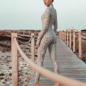 Może przedstawiać: Kombinezon z nadrukiem wężowej skóry w odcieniach szarości i bieli. Strój ma wysoki dekolt i długie rękawy. Osoba ma na sobie przezroczyste sandały na obcasie. Tło obejmuje drewnianą promenadę i plażę.