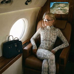 Op de afbeelding: Een persoon in een jumpsuit met slangenprint en een bril zit in een privéjet. Een zwarte handtas staat op een houten richel bij een rond raam. Het interieur heeft bruine leren stoelen en houten panelen.