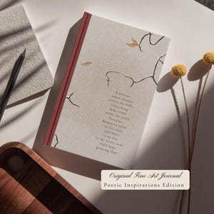 May include: A hardcover journal with a light gray cover featuring a dark red spine, gold foil branch illustrations, and the text: A delicate white blanket covers the hills. Everything falls silent while the earth breathes. Memories seem so far away and there is no sun in my weary soul. Only hope growing slow.