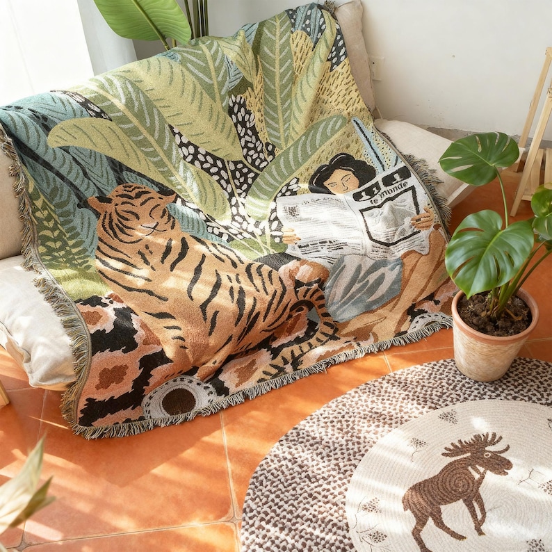 May include: A woven throw blanket featuring a tiger, tropical leaves, and a person reading a newspaper. The blanket has a fringed edge and a color palette of green, brown, and beige. The newspaper reads "le monde".