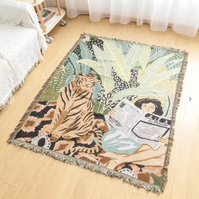 May include: Woven throw blanket with a jungle scene featuring a tiger, lush foliage, and a person reading a newspaper. The blanket has a fringed edge and a color palette of beige, green, and brown.
