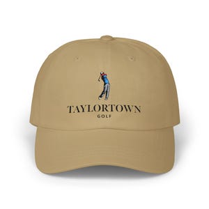 May include: A tan baseball cap featuring the text "TAYLORTOWN GOLF" in black, with a graphic of a golfer in action. The cap has a curved brim and a structured crown, suitable for outdoor activities.
