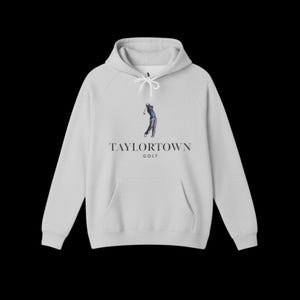 Könnte beinhalten: Ein hellgrauer Kapuzenpullover mit einer Vordertasche und einer Kapuze mit Kordelzug. Auf der Vorderseite steht der Text "TAYLORTOWN GOLF" mit einer Illustration eines Golfspielers im Schwung. Das Wort "GOLF" steht darunter.