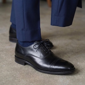 May include: Black leather Oxford shoes with a cap toe and lace-up fastening. The shoes are worn with navy blue socks and trousers. The shoes have a polished finish, suitable for formal wear.