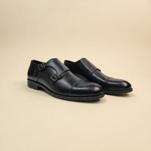 Puede incluir: Un par de zapatos negros de cuero con doble hebilla. Los zapatos tienen un dise&ntilde;o de puntera y dos hebillas ajustables. Se muestran sobre un fondo neutro.