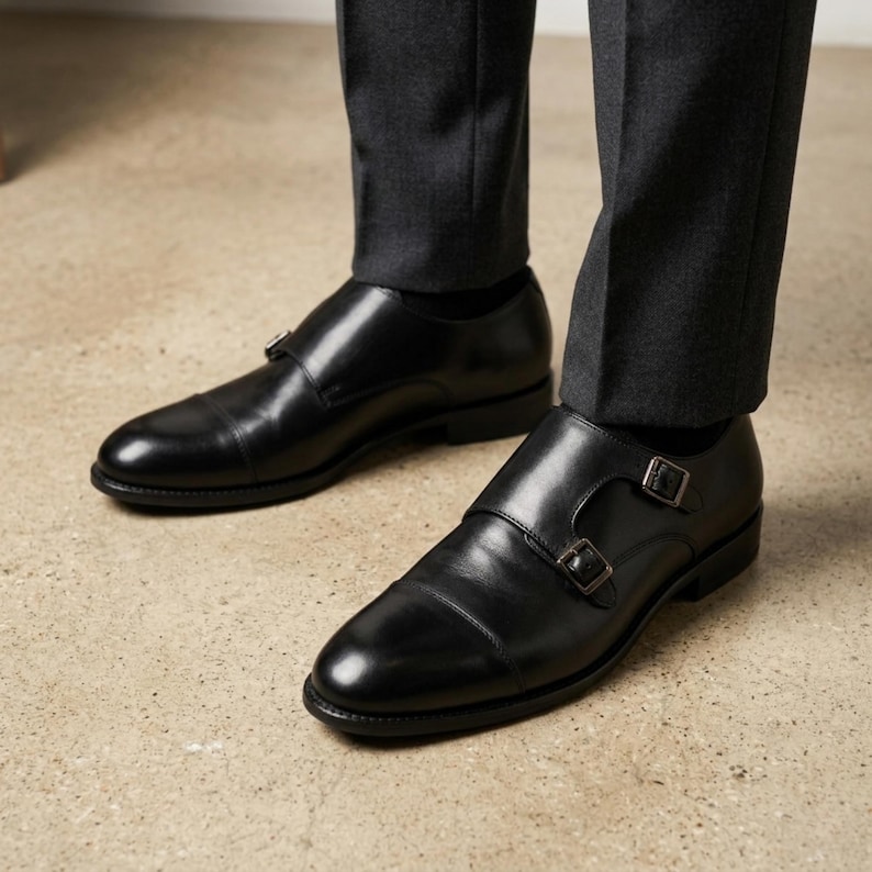 Puede incluir: Zapatos negros de cuero con doble hebilla y hebillas plateadas. Los zapatos tienen un dise&ntilde;o de puntera y se usan con pantalones de vestir grises oscuros. Los zapatos est&aacute;n sobre una superficie marr&oacute;n claro.