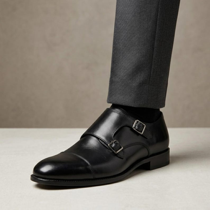 Puede incluir: Zapatos negros de cuero con doble hebilla y hebillas plateadas. Los zapatos tienen un dise&ntilde;o de puntera y se usan con pantalones grises oscuros. La imagen muestra un primer plano de los zapatos y parte de las piernas.