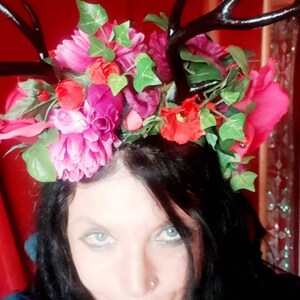 Op de afbeelding: Een vrouw met donker haar en blauwe ogen draagt een bloemenkrans met zwarte geweien. De krans is versierd met roze, rode en groene bloemen.