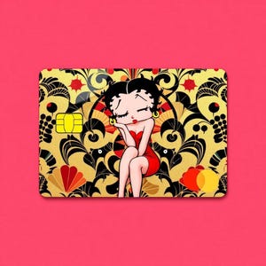May include: A credit card featuring a cartoon image of Betty Boop in a red dress, resting her chin on her hand. The card has a gold and black floral design background and a pink background.