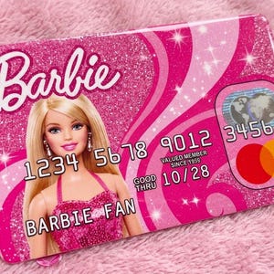 K&ouml;nnte beinhalten: Ein rechteckiger, pinkfarbener Kreditkartenaufkleber mit Glitzereffekt und Barbie-Logo. Das Design zeigt eine blonde Puppe in einem pinken Kleid, den Text BARBIE FAN, die Nummer 1234 5678 9012 3456, das Datum GOOD THRU 10/28 und ein rundes Logo.