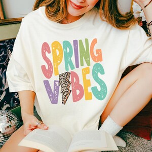 May include: Cream-colored t-shirt with the words "SPRING VIBES" in colorful, playful lettering. The design includes a lightning bolt with a leopard print pattern. The shirt is a casual, comfortable style.
