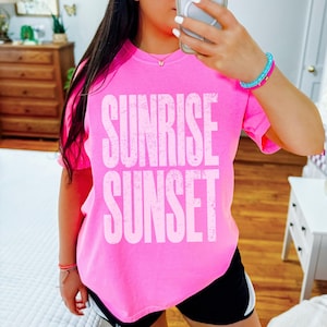 May include: Bright pink t-shirt with the words "SUNRISE SUNSET" in large, white, distressed lettering. The shirt is a vibrant color and appears to be made of soft material. The overall style is casual and relaxed.
