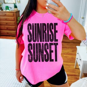 May include: Bright pink t-shirt with the words "SUNRISE SUNSET" in large, distressed black letters. The shirt is a vibrant color and appears to be made of soft material. The overall style is casual and comfortable.