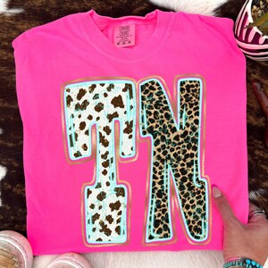 Puede incluir: Una camiseta rosa brillante con las letras "TN" en un diseño audaz. La "T" presenta un estampado de piel de vaca, mientras que la "N" tiene un estampado de leopardo. Ambas letras están delineadas en azul claro y dorado. La camiseta tiene cuello redondo y mangas cortas.
