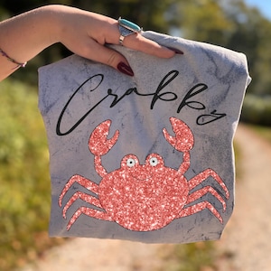 Op de afbeelding: Lichtgrijs t-shirt met een rode glitterkrabontwerp en het woord "Crabby" in zwart script. De krab heeft een lachend gezicht. De achtergrond is een vage buitenscène.