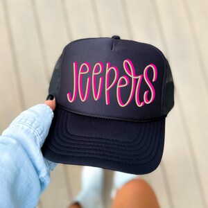 May include: A navy blue trucker hat with a mesh back and a curved brim. The word "JEEPERS" is written across the front in a light green font with a bright pink outline. The hat has a black rope detail along the brim.