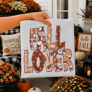 May include: A light gray sweatshirt featuring the text FALL LOVER in large, blocky letters. The letters are filled with various brown and tan patterns, including plaid, polka dots, pumpkins, pinecones, and leaves, with a stitched-style outline.