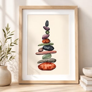 May include: A framed print featuring a watercolor painting of a balanced stack of colorful stones. The stones vary in size and color, including red, green, purple, and black. The artwork is set against a cream-colored background and framed in light wood.