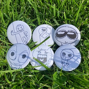 May include: Six white round buttons with black line art illustrations of various cartoon characters. The buttons are arranged on a bed of green grass. Each button features a unique, expressive face or figure.