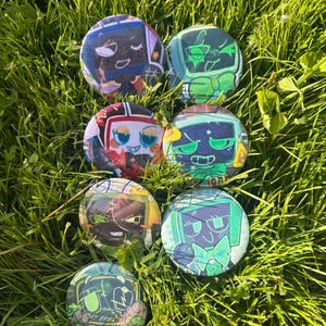 May include: Seven circular pin-back buttons featuring stylized character illustrations. The designs include various figures with television-shaped heads, vibrant color palettes of green, purple, and red, and expressive facial features in a graphic art style.