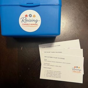Puede incluir: Una caja de plástico azul con una pegatina circular blanca que dice "Raising Capable Humans". Junto a la caja hay tres tarjetas blancas con instrucciones. Las tarjetas incluyen el texto "Do Laundry Start to Finish."