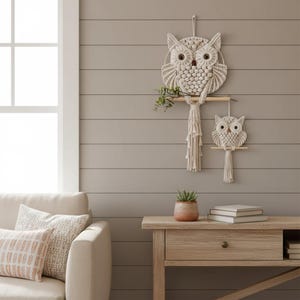 Könnte beinhalten: Zwei Wandbehänge in Form von Eulen aus Makramee. Die größere Eule hat einen kreisförmigen Rahmen und sitzt auf einer Holzstange mit Grünzeug. Die kleinere Eule befindet sich darunter. Beide bestehen aus cremefarbenem Seil und hängen an einer grauen Wand.