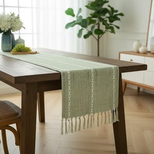 Könnte beinhalten: Ein salbeigrüner Tischläufer mit weißen Stickdetails und Fransen. Der rechteckige Läufer wird auf einem dunklen Holztisch präsentiert. Der Läufer hat vertikale weiße Stickmuster und horizontale weiße Streifen.