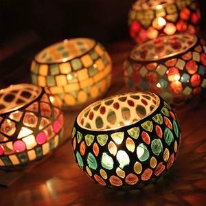 May include: A collection of colorful mosaic glass candle holders. The holders are round with a variety of colors including yellow, orange, red, green, and blue. Each holder has a candle lit inside, creating a warm glow.