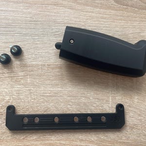 Può includere: Componenti in plastica nera, tra cui un pezzo curvo con maniglia, un pezzo rettangolare con magneti incorporati e due piccole viti. Le parti sono disposte su una superficie di legno chiaro.
