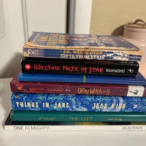 Puede incluir: Una pila de siete libros con varias portadas. Los títulos incluyen "Dr. Wayne Dyer Gifts From Eykis", "Witches' Night of Fear" y "Things in Jars". Los libros son de diferentes colores, incluyendo azul, verde y rojo, apilados sobre una superficie blanca.