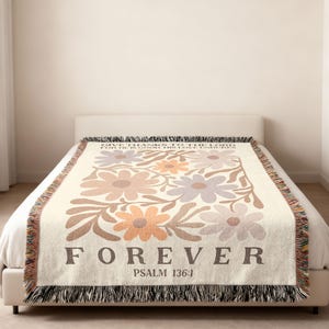 Puede incluir: Manta de color crema con un diseño floral en tonos marrones, naranjas y morados. La palabra "FOREVER" está impresa en letras grandes de color marrón, con "SALMO 136:1" debajo. La manta tiene un borde con flecos.