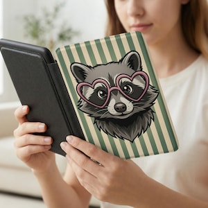May include: A black e-reader case with a cover featuring a raccoon wearing pink heart-shaped glasses. The raccoon is set against a background of green and beige vertical stripes. The e-reader is held open.