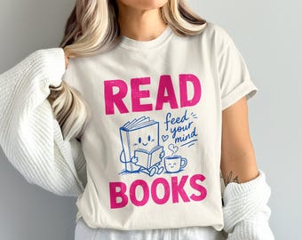 Op de afbeelding: Een crèmekleurig t-shirt met korte mouwen en de tekst READ BOOKS in grote roze letters. Tussen de woorden staat een blauwe afbeelding van een lachend boek dat een kleiner boek leest naast een lachende koffiemok met de tekst feed your mind.