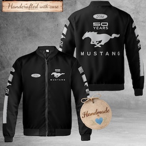 Op de afbeelding: Zwarte bomberjack met het Ford Mustang logo en tekst. De jas heeft een ritssluiting en witte accenten op de mouwen. De achterkant toont het Mustang logo en de tekst "50 YEARS". De jas is gelabeld "Handcrafted with care".