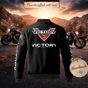 以下が含まれることがあります： 背中と袖に白と赤の「Victory Motorcycles」ロゴが入った黒いボンバージャケット。背景には2台のバイクがあり、上部には「Handcrafted with care」の文字があります。