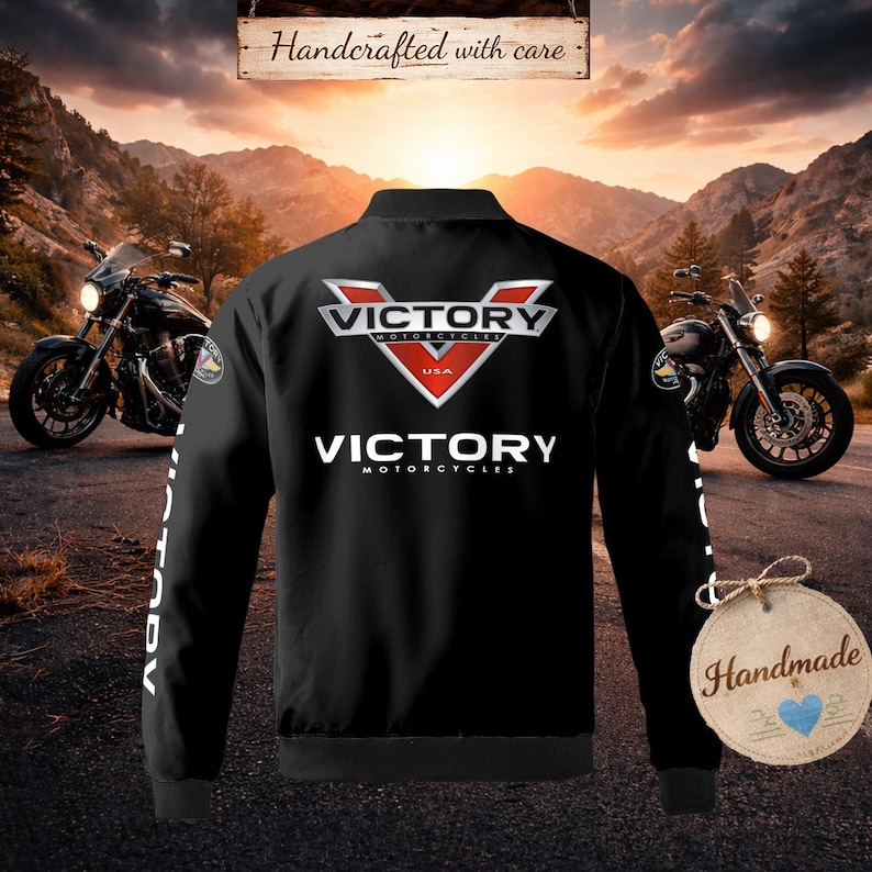 以下が含まれることがあります： 背中と袖に白と赤の「Victory Motorcycles」ロゴが入った黒いボンバージャケット。背景には2台のバイクがあり、上部には「Handcrafted with care」の文字があります。